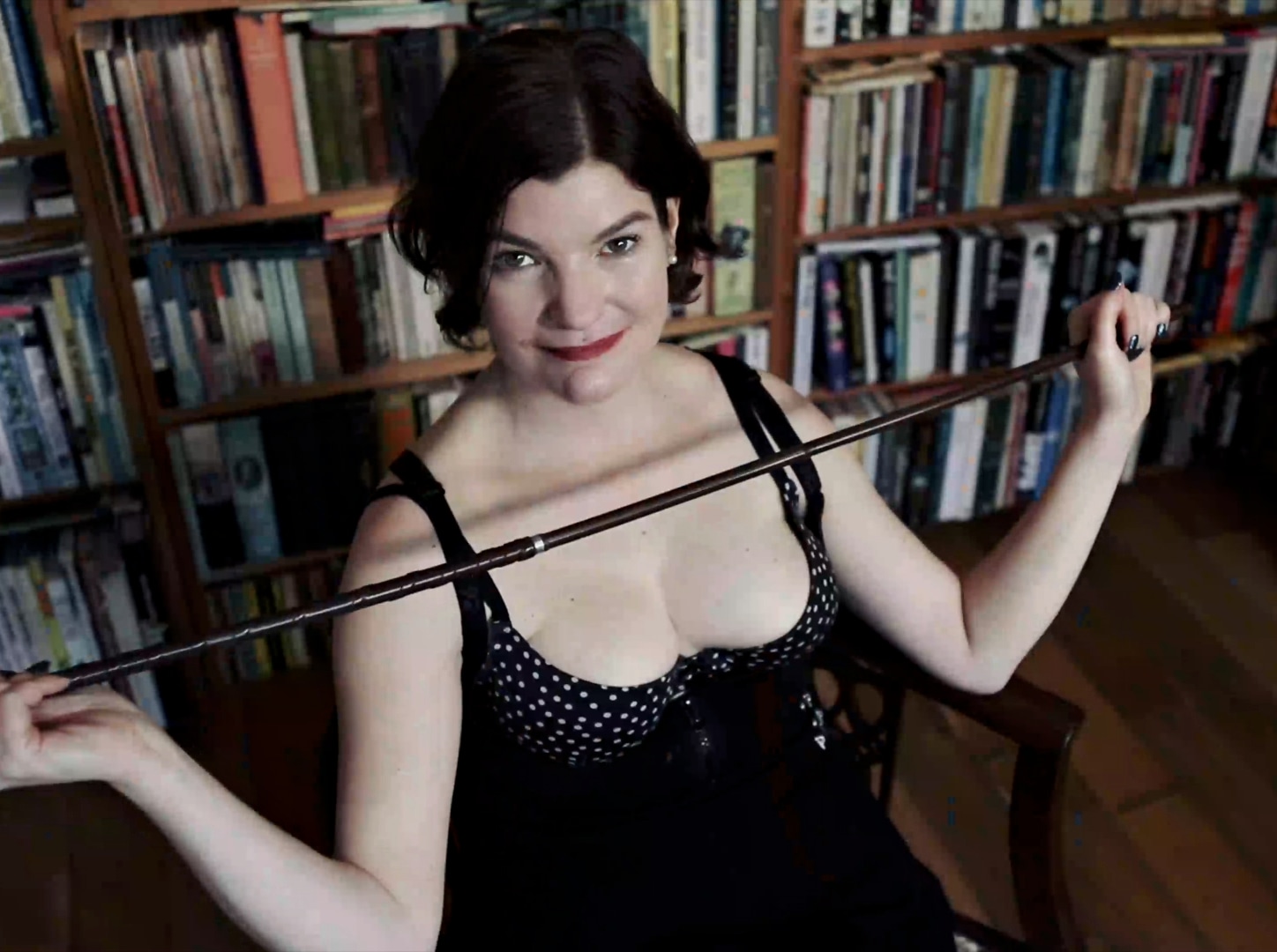 Miss Matthews, wearing a corset, seated in a chair flexing a menacing cane.